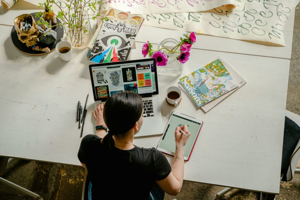 Mujer diseñadora trabajando en una mesa blanca con un portátil y una tablet, rodeada de revistas, flores, café y herramientas de dibujo.